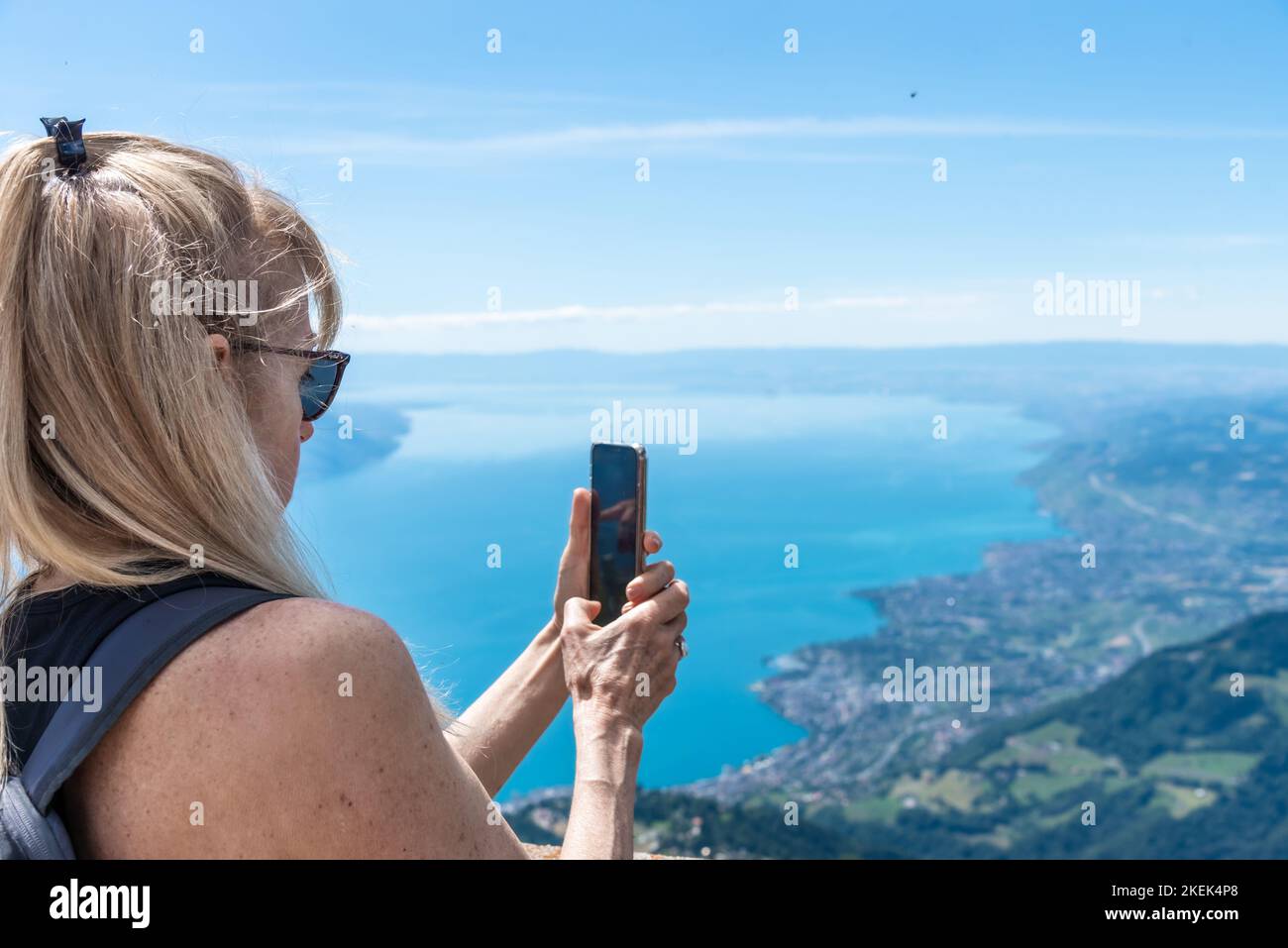 Donna bionda matura con occhiali da sole e zaino che scatta una foto del lago di Ginevra da una montagna. Foto Stock