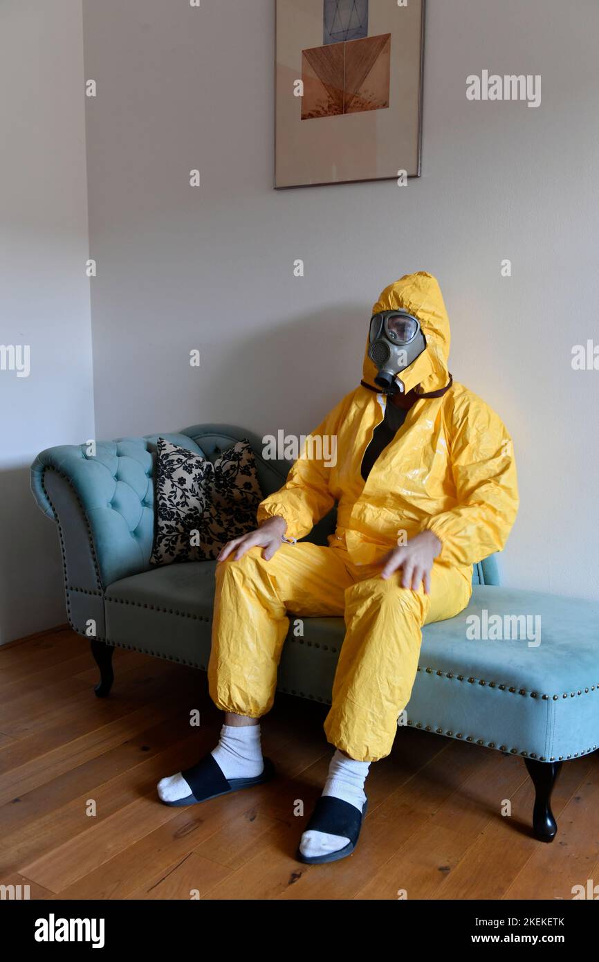 una persona in tuta gialla e maschera a gas seduta sul divano a casa Foto Stock