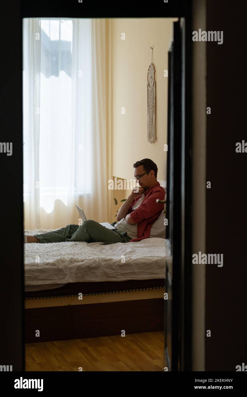 Uno studente giovane e penoso si siede sul letto con un computer portatile in ginocchio guardando un video didattico dall'università Foto Stock