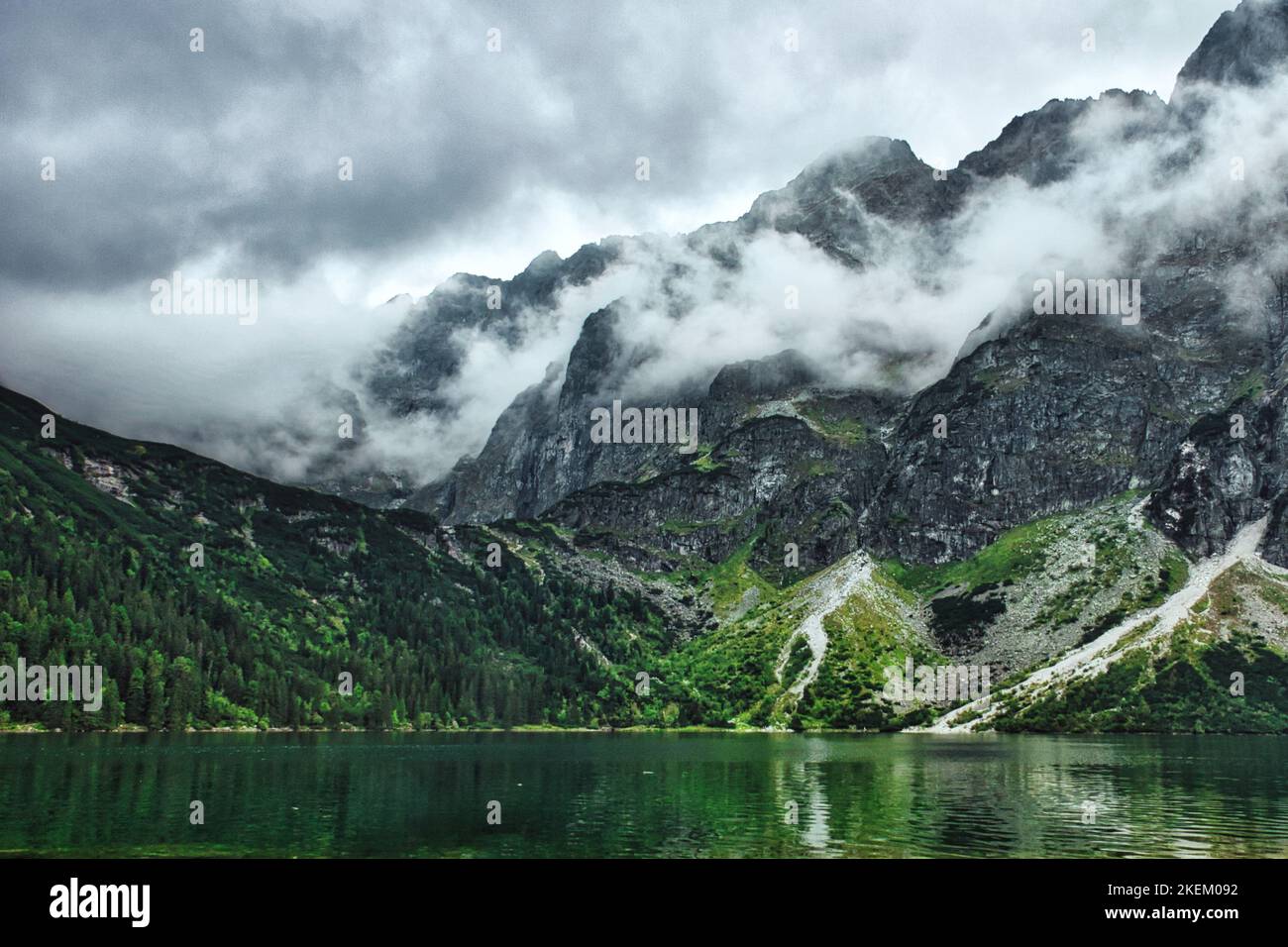 Nuvole drammatiche sopra le scogliere di picco di Rysy, foresta che si riflette nel lago di montagna Morskie Oko nel parco nazionale di Tatra, in Polonia, vicino a Zakopane Foto Stock