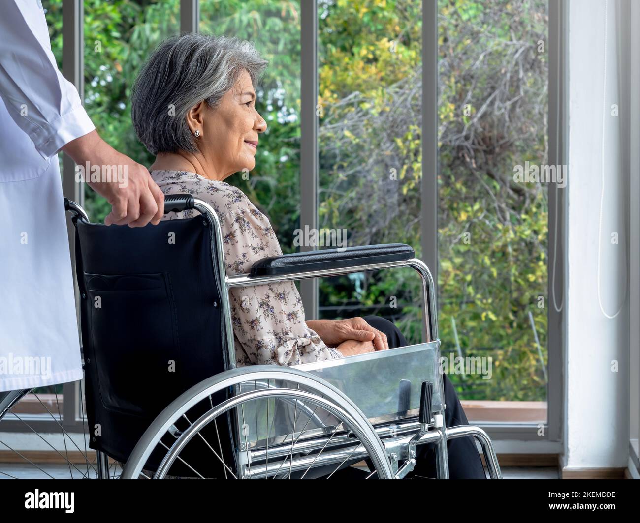 Primo piano ritratto di felice asiatico anziano paziente femminile si siede in sedia a rotelle, mentre uomo medico in camice bianco prendersi cura in ufficio medico a hospit Foto Stock