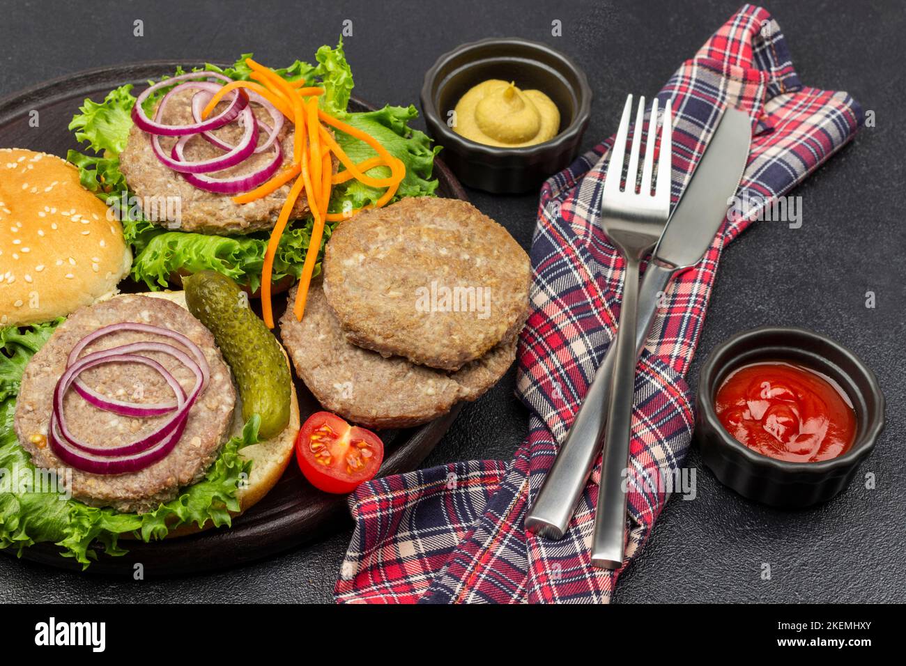 Salsa di pomodoro e senape in ciotole. Coltello e forchetta sul tovagliolo. Panini rotondi, hamburger fritto, sottaceti, cipolla e lattuga sul piatto. Sfondo nero. Foto Stock