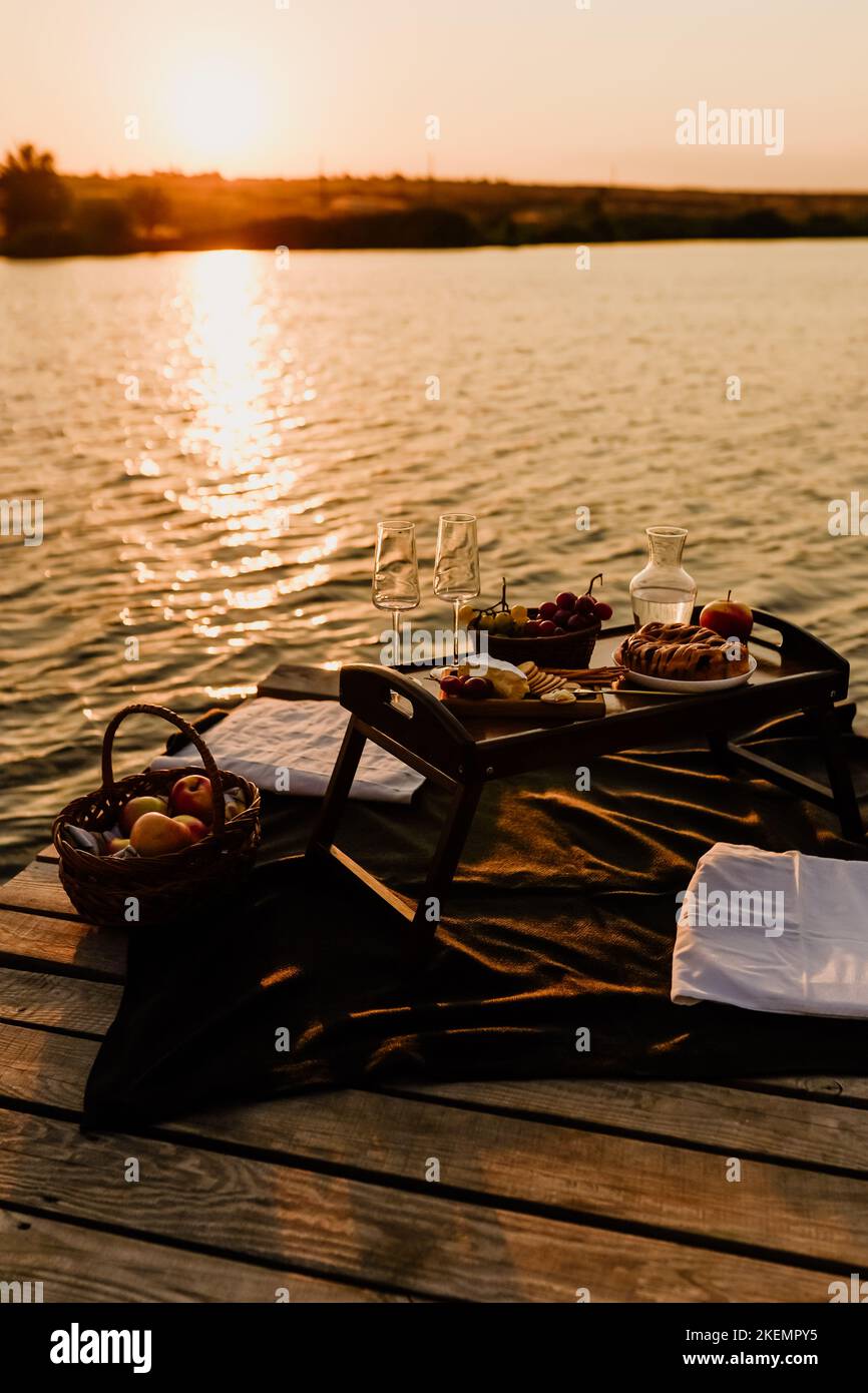 Incredibile tramonto sul lago, vino bianco, torta di frutta, mele, uva, formaggio brie, biscotti. Picnic estetico autunnale. Foto Stock