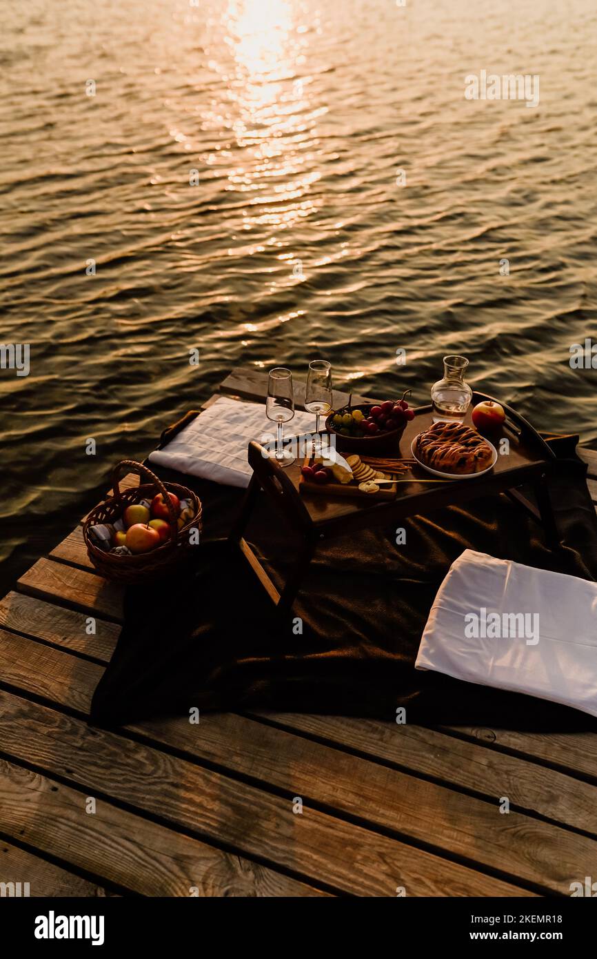 Incredibile tramonto sul lago, vino bianco, torta di frutta, mele, uva, formaggio brie, biscotti. Picnic estetico autunnale. Foto Stock