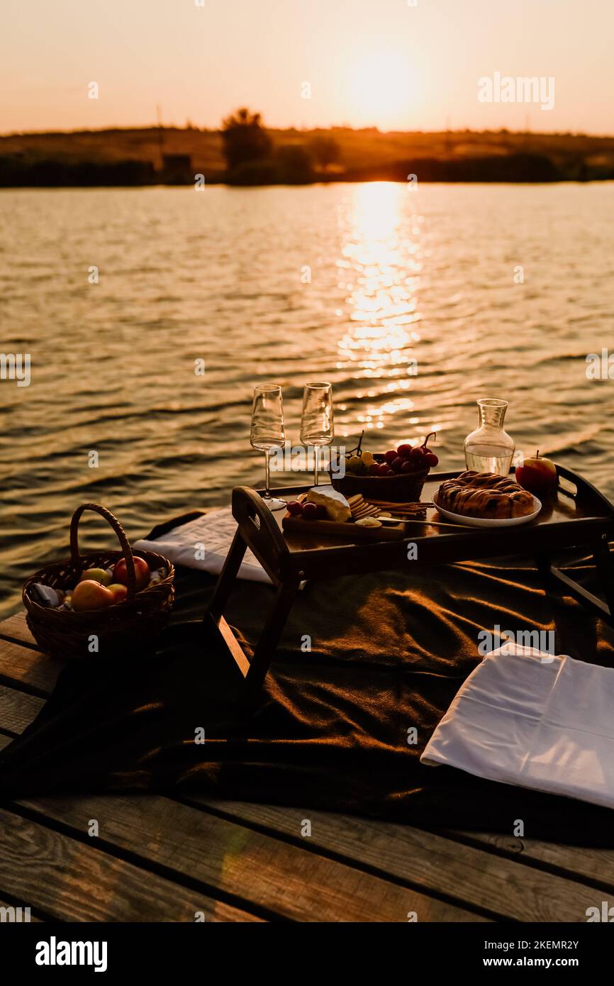 Incredibile tramonto sul lago, vino bianco, torta di frutta, mele, uva, formaggio brie, biscotti. Picnic estetico autunnale. Foto Stock