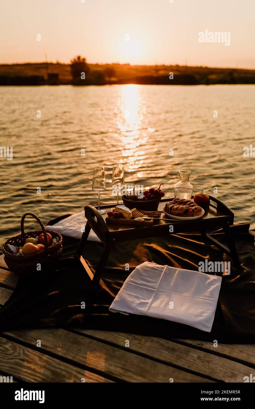 Incredibile tramonto sul lago, vino bianco, torta di frutta, mele, uva, formaggio brie, biscotti. Picnic estetico autunnale. Foto Stock