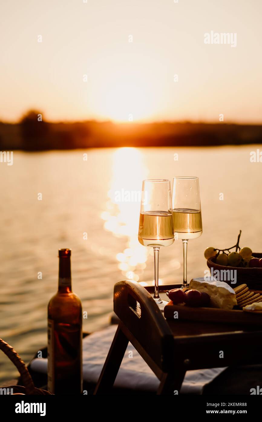 Incredibile tramonto sul lago, vino bianco, torta di frutta, mele, uva, formaggio brie, biscotti. Picnic estetico autunnale. Foto Stock