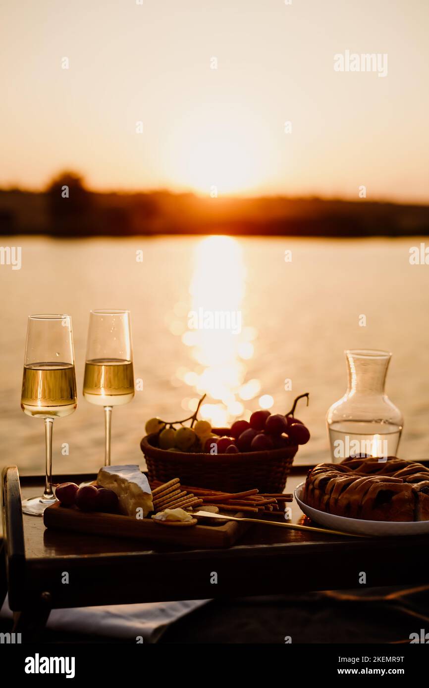 Incredibile tramonto sul lago, vino bianco, torta di frutta, mele, uva, formaggio brie, biscotti. Picnic estetico autunnale. Foto Stock