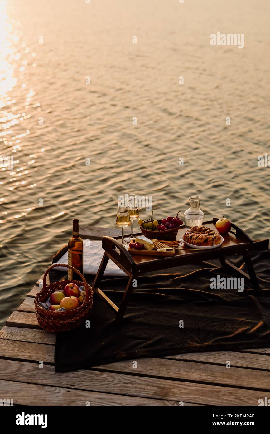 Incredibile tramonto sul lago, vino bianco, torta di frutta, mele, uva, formaggio brie, biscotti. Picnic estetico autunnale. Foto Stock
