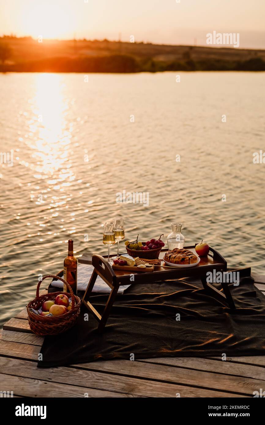 Incredibile tramonto sul lago, vino bianco, torta di frutta, mele, uva, formaggio brie, biscotti. Picnic estetico autunnale. Foto Stock