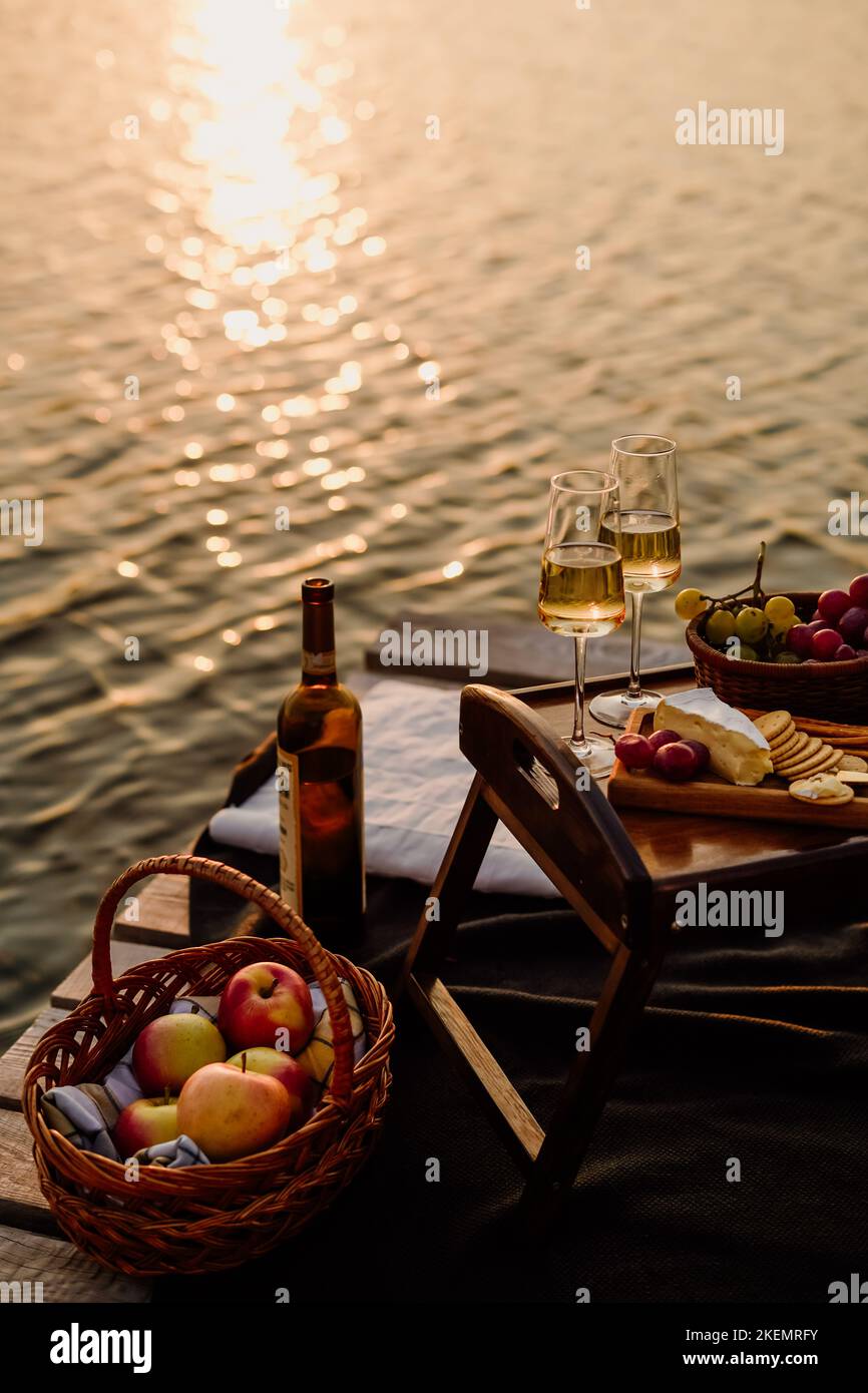 Incredibile tramonto sul lago, vino bianco, torta di frutta, mele, uva, formaggio brie, biscotti. Picnic estetico autunnale. Foto Stock
