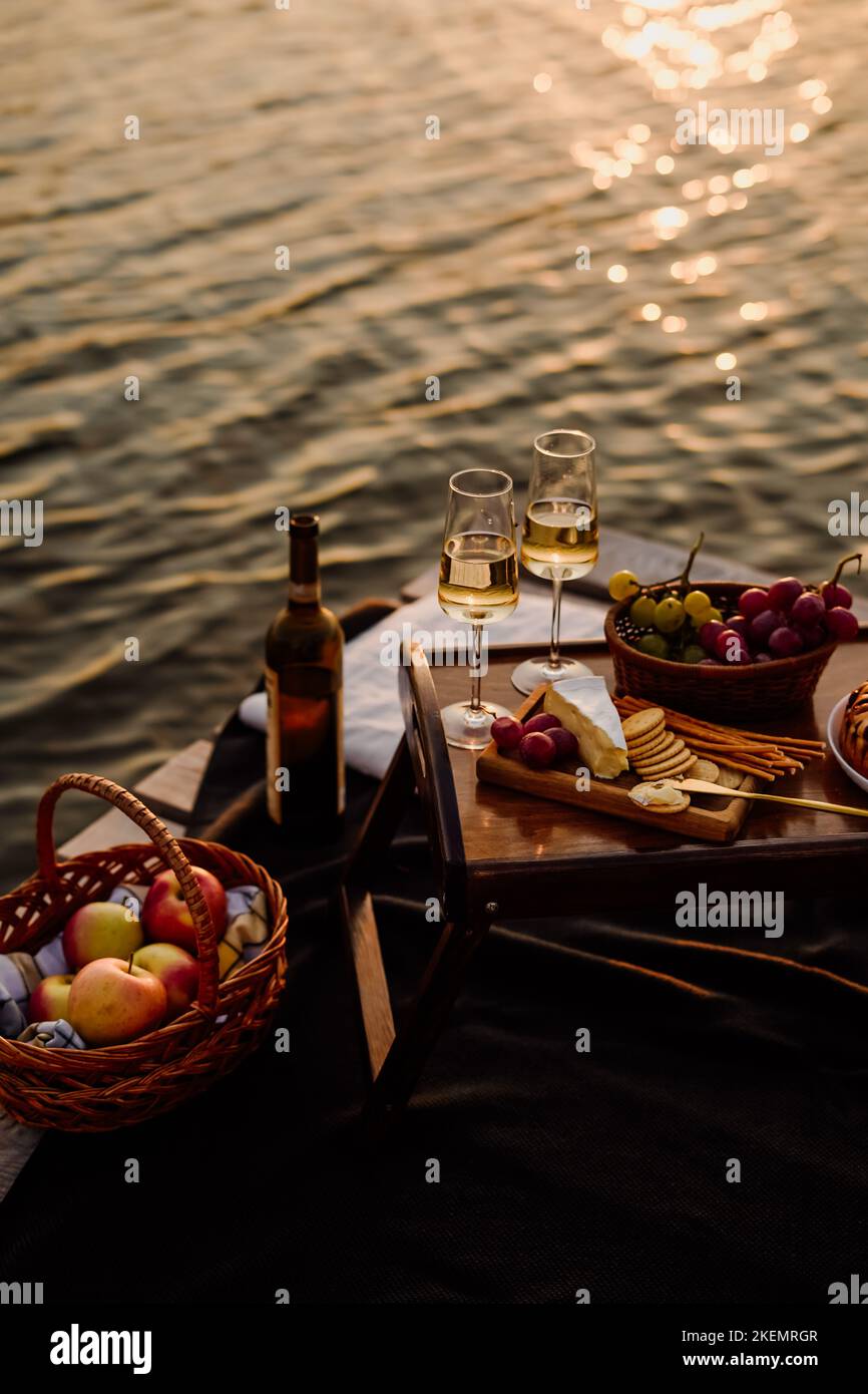 Incredibile tramonto sul lago, vino bianco, torta di frutta, mele, uva, formaggio brie, biscotti. Picnic estetico autunnale. Foto Stock