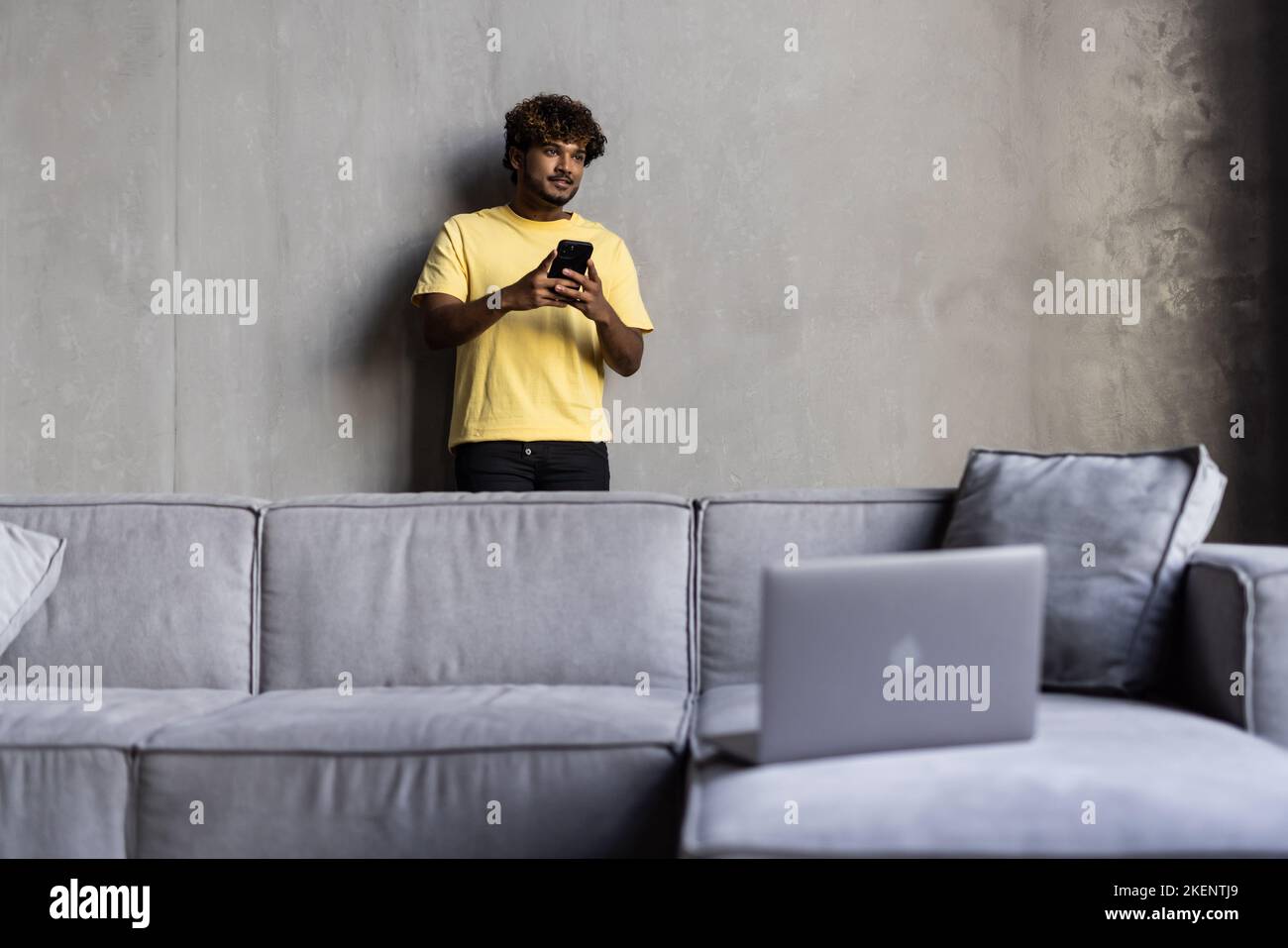 Un bel ragazzo indiano o arabo, sedersi in un moderno soggiorno su un comodo divano, utilizza smartphone, messaggi di testo, navigare sui social media, sur Foto Stock