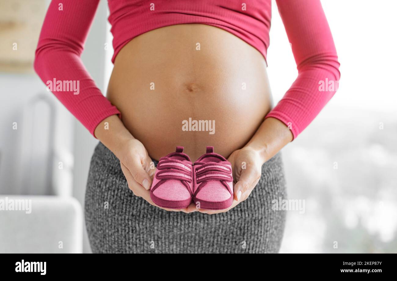 Gravidanza. Donna incinta in attesa bambina che tiene scarpe rosa rosso in rivelazione di sesso. Attesa mamma che mostra il pancia del bambino. Abbigliamento per bambini Foto Stock