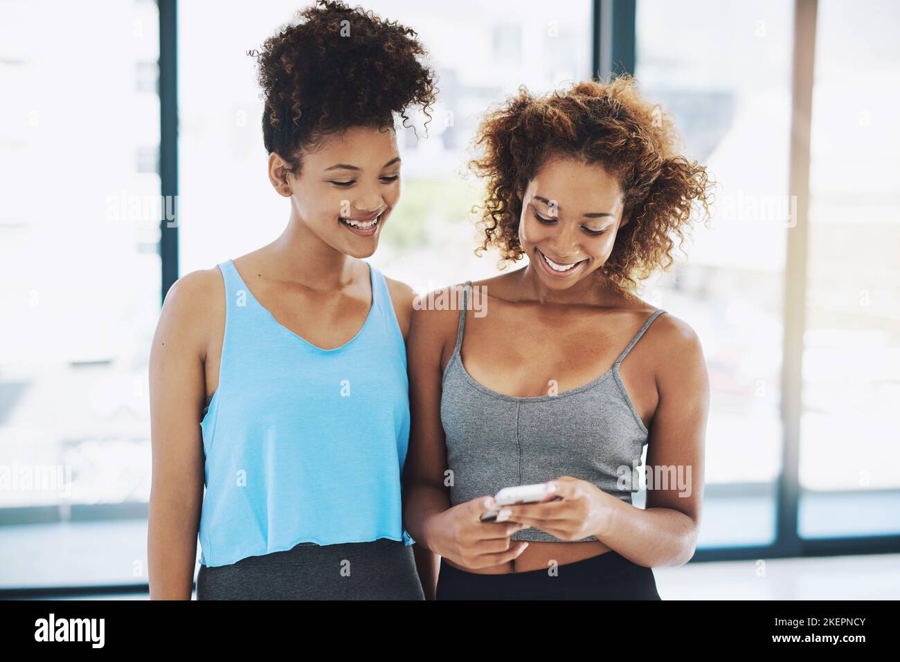 Raccontiamo a tutti la nostra sessione di allenamento. Due giovani donne in forma che chiacchierano mentre guardano un telefono all'interno di una palestra prima di una sessione di allenamento. Foto Stock
