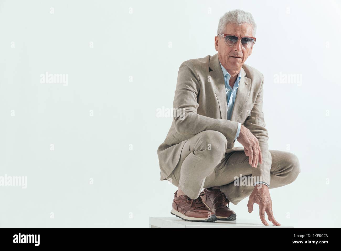 l'uomo anziano con gli occhiali da sole che sono freschi, accovacciati e tenendo le braccia in posa di moda di fronte allo sfondo grigio Foto Stock