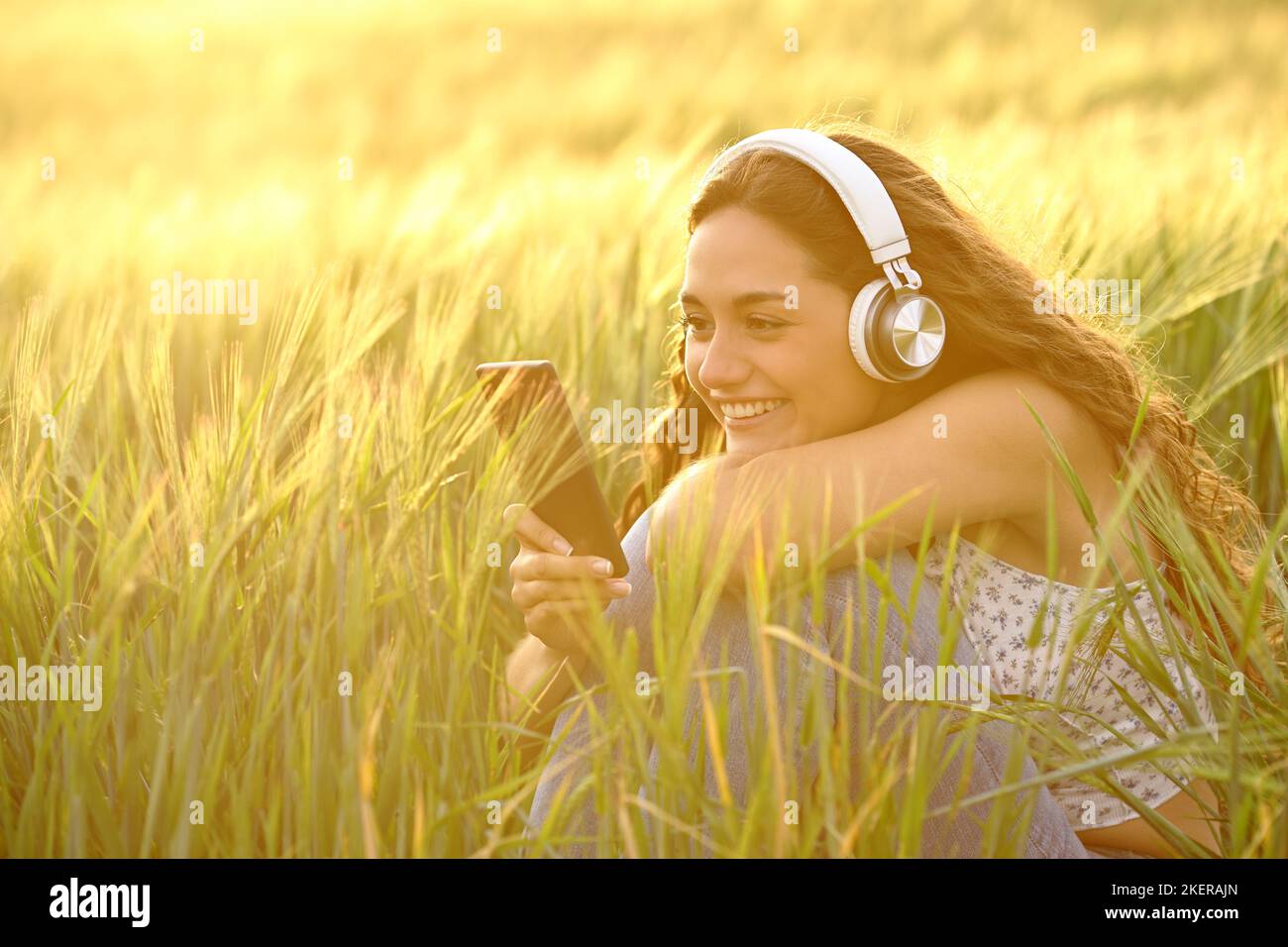 Donna felice al tramonto indossando cuffie che ascoltano musica da smartphone in un campo di grano Foto Stock
