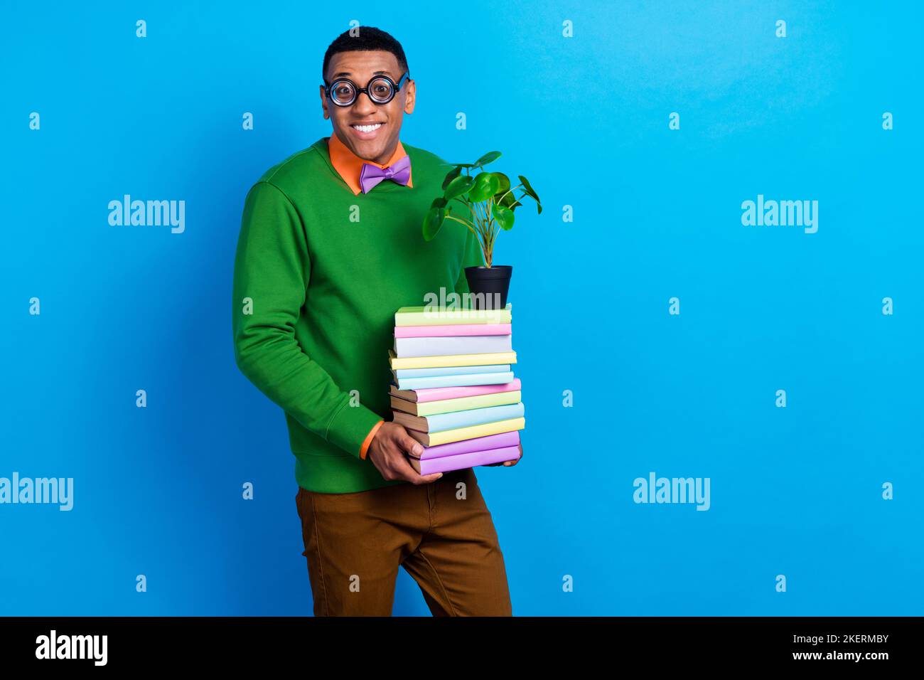 Foto di giovane simpatico sorridente studente accademico nerd indossare occhiali funky tenere libri stack con piante decorative isolato su sfondo di colore blu Foto Stock