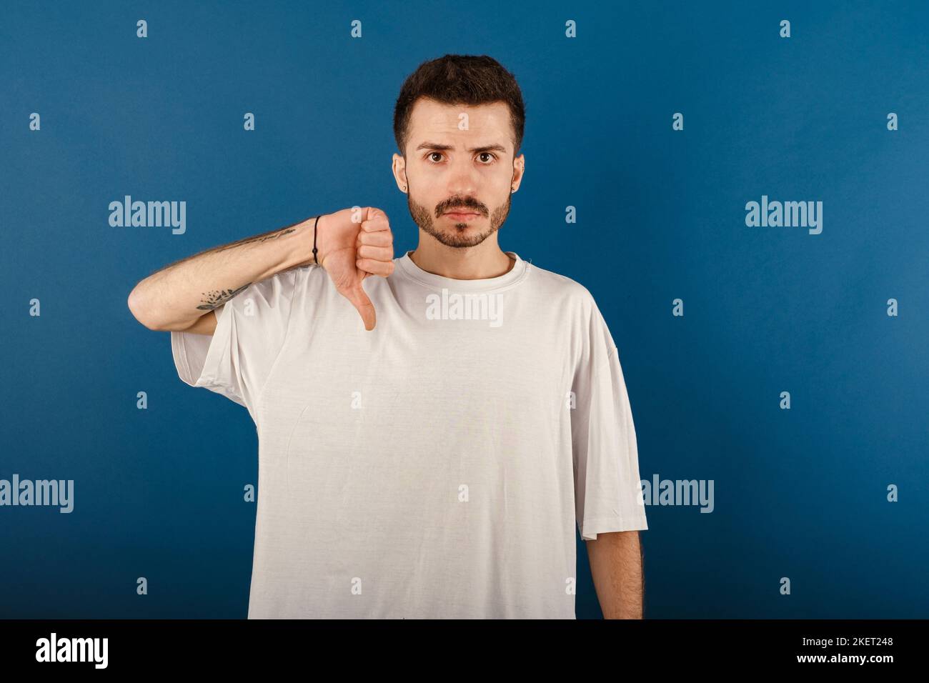 Uomo caucasico che indossa un tee bianco che posa isolato su sfondo blu mostrando il pollice verso il basso ed esprimendo dispiacere. Grumpy, insoddisfatto, infastidito, infelice Foto Stock