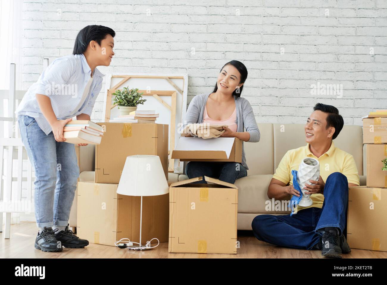 Amorevole famiglia asiatica chiacchierando animatedly con l'altro mentre disimballando roba in spazioso soggiorno di nuovo appartamento, mucchi di scatole in movimento Foto Stock