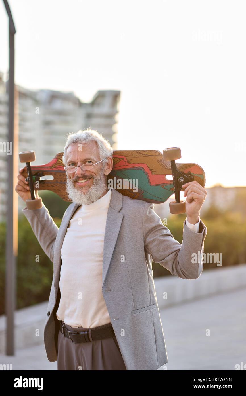 Attivo felice cool vecchio skater uomo d'affari che tiene skateboard in città. Foto Stock