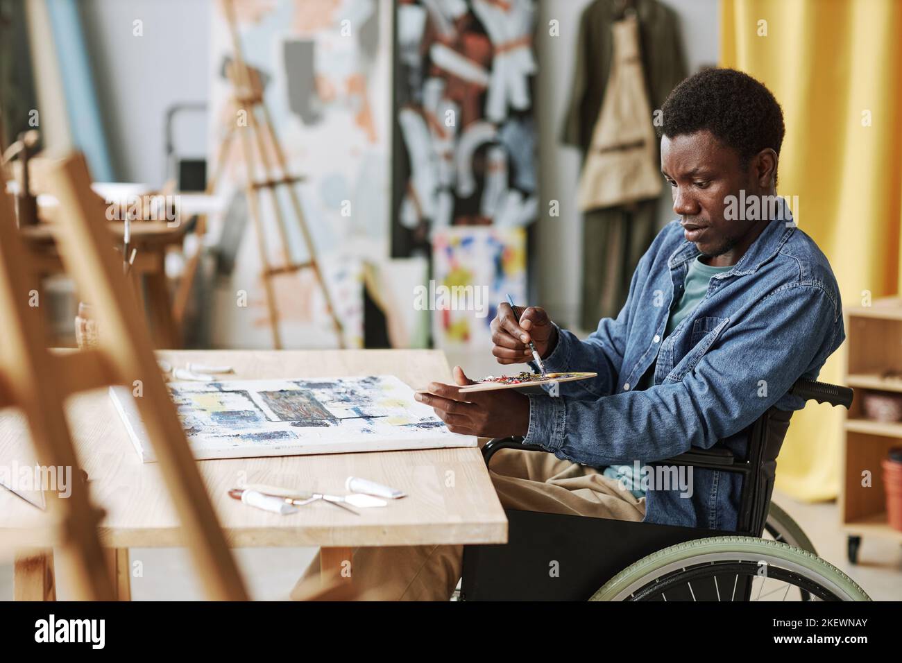 Giovane artigiano afroamericano in sedia a rotelle che mescola i colori sulla tavolozza mentre si siede al tavolo in studio di arte e pittura su tela Foto Stock