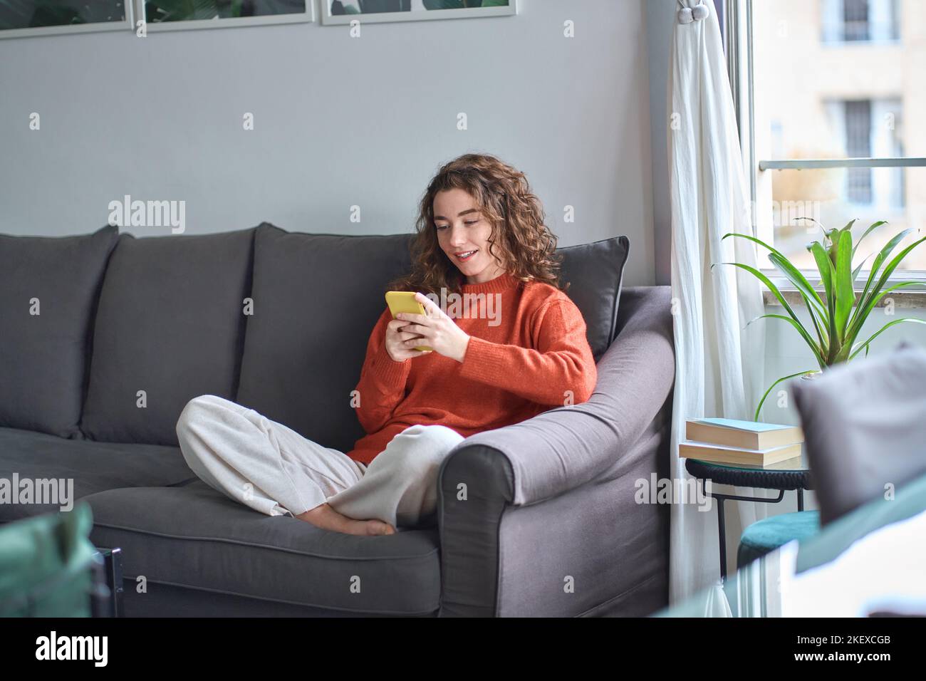 Giovane donna sorridente seduta sul divano utilizzando il telefono cellulare a casa. Foto Stock