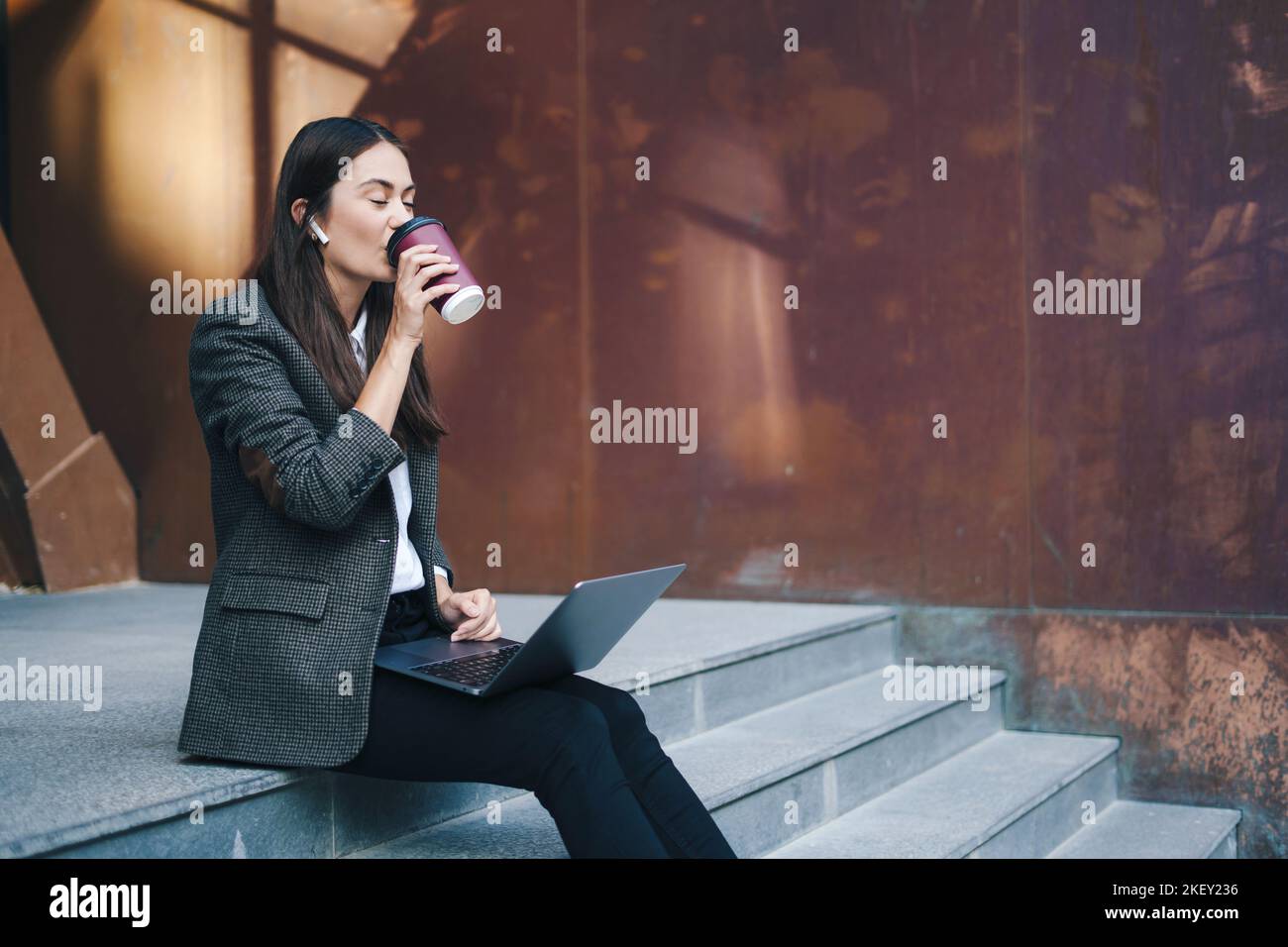 Giovane e attraente donna d'affari che beve caffè e lavora con il computer portatile mentre si siede sulle scale all'aperto. Lavoro online, business online. Femmina Foto Stock