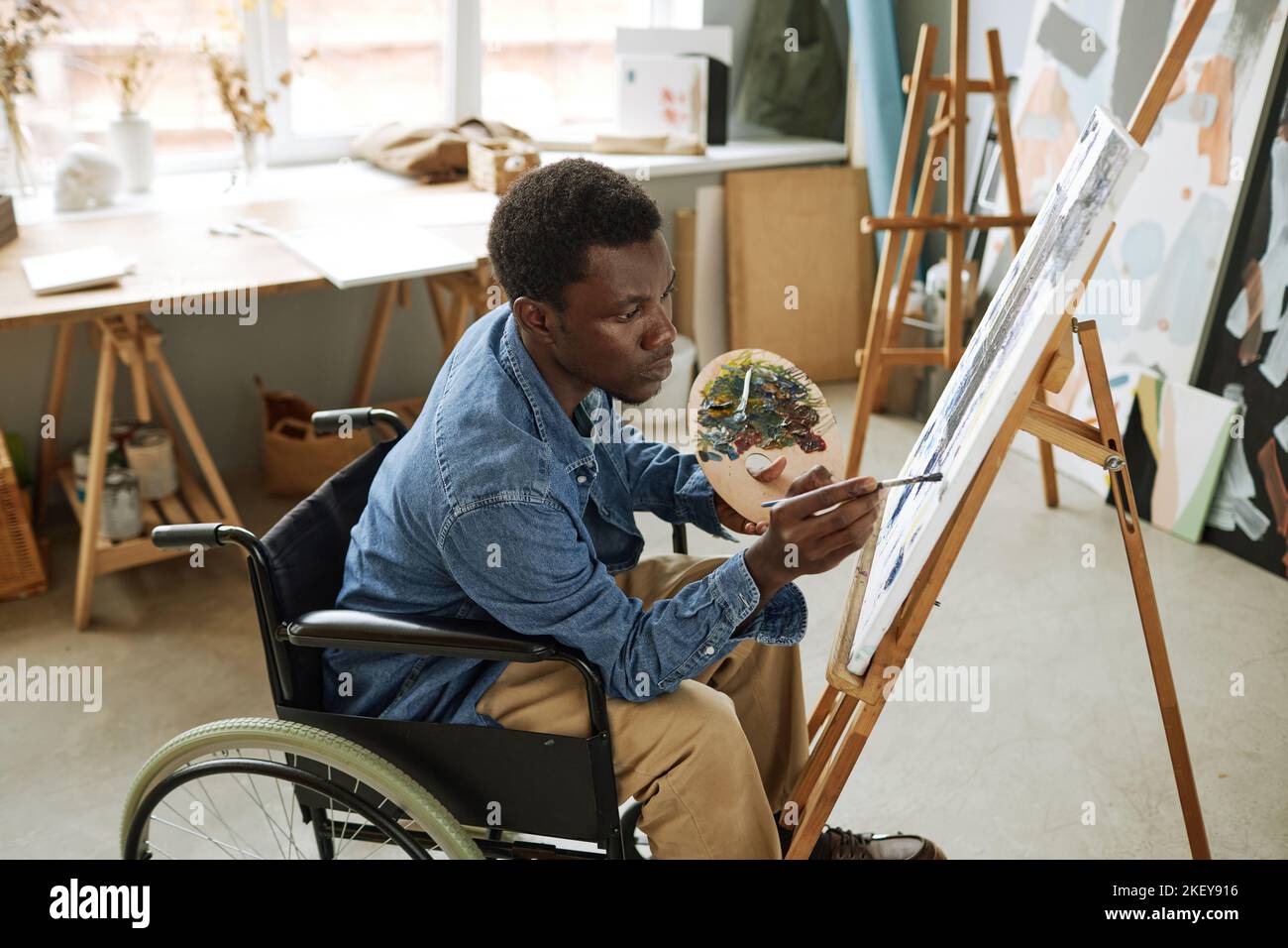 Giovane uomo nero di talento con tavolozza di colori che dipinge un nuovo capolavoro in studio d'arte mentre si siede in sedia a rotelle di fronte al cavalletto Foto Stock