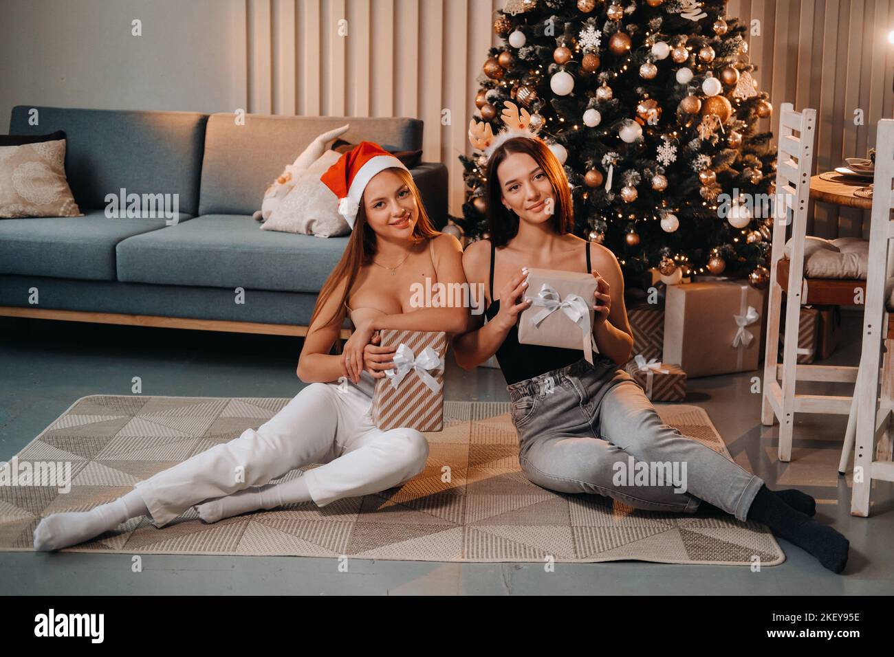 Due belle ragazze giovani con i regali di Natale nelle loro mani sono seduti vicino all'albero di Natale nel house.friends con gli abiti di Natale e. Foto Stock