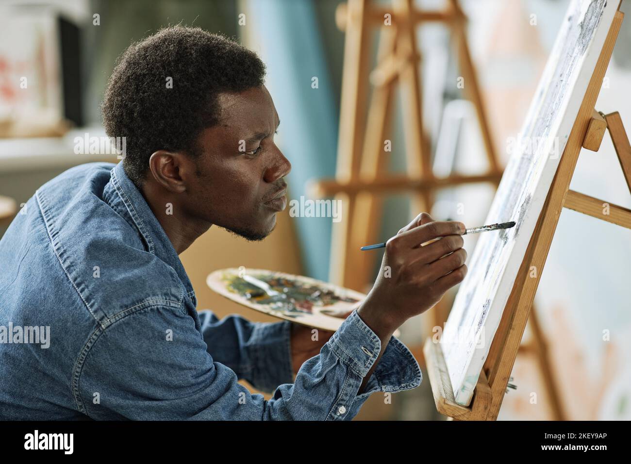 Vista laterale di un giovane uomo nero serio in camicia denim dipingere nuove opere d'arte con vernici acriliche mentre si siede davanti al cavalletto con tela Foto Stock