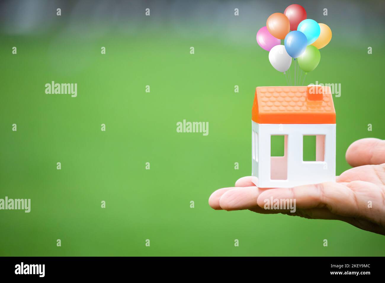 Modello di casa attaccato a palloncini colorati isolato su sfondo verde. Figura di casa di tenuta della mano. Significato metaforico delle opportunità perse. Foto Stock