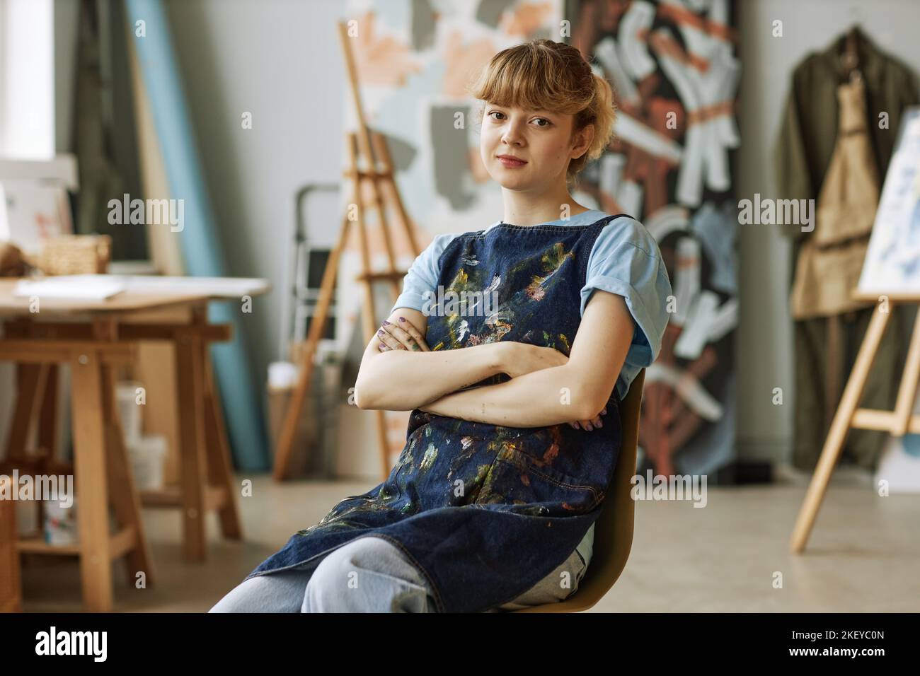 Giovane bionda artista o insegnante di pittura seduto su sedia in studio o in aula di scuola d'arte con le braccia incrociate sul petto Foto Stock
