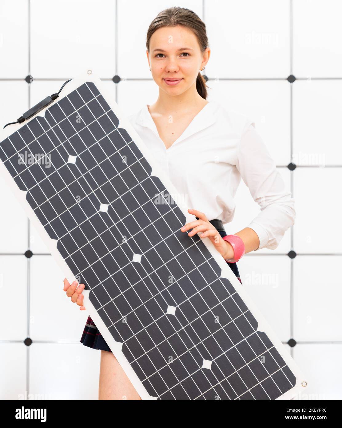 Una giovane donna su un fondo industriale bianco tiene una cella fotovoltaica flessibile a batteria solare nelle sue mani Foto Stock