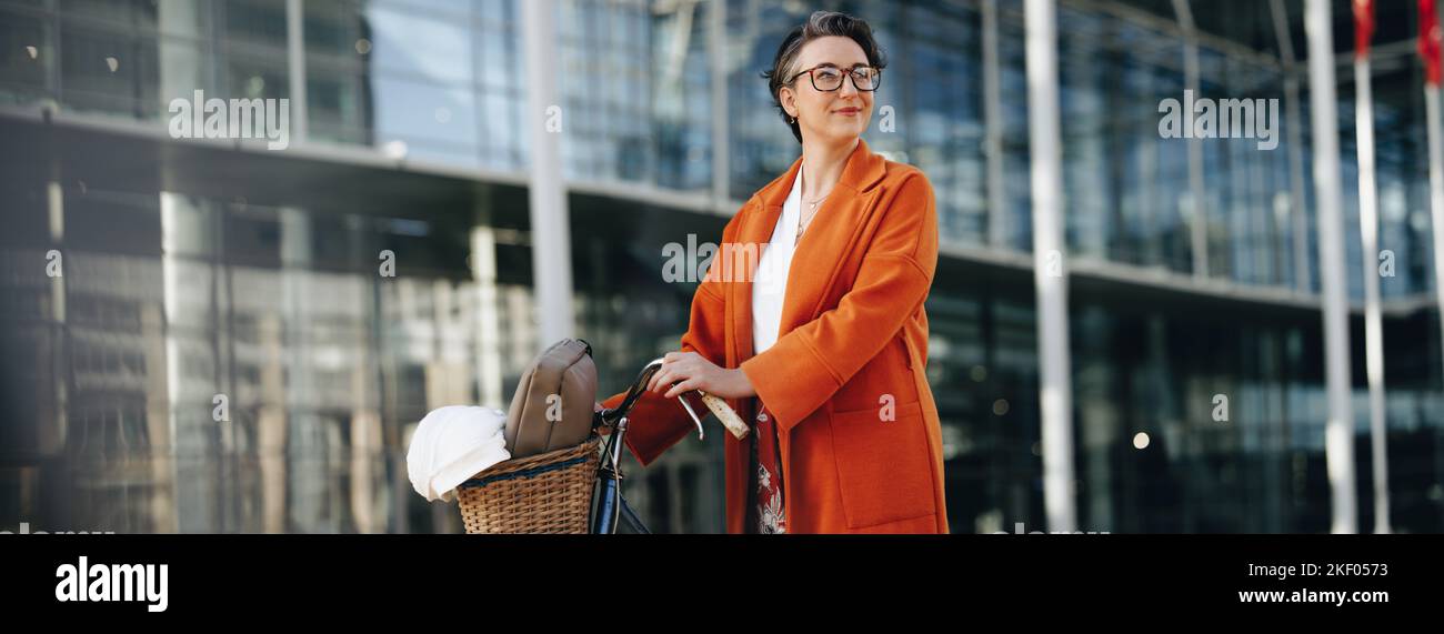 Donna d'affari premurosa che spinge la sua bici durante il suo viaggio mattutino in città. Donna d'affari matura guardando via pensivamente mentre camminando per lavorare w Foto Stock