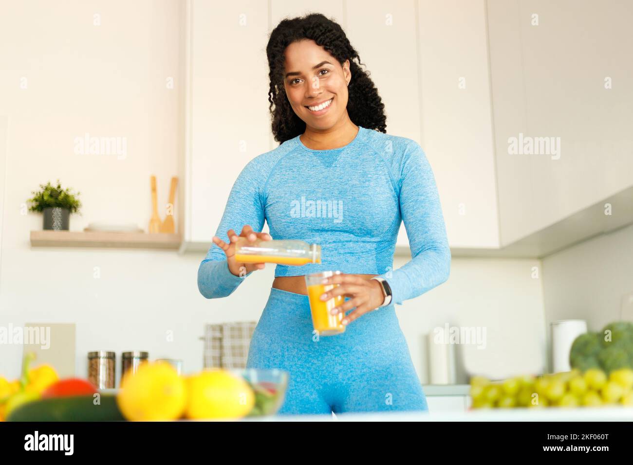 Donna in buona forma nera gustando un pranzo gustoso e versando succo d'arancia dalla brocca al bicchiere, in piedi in cucina a casa Foto Stock
