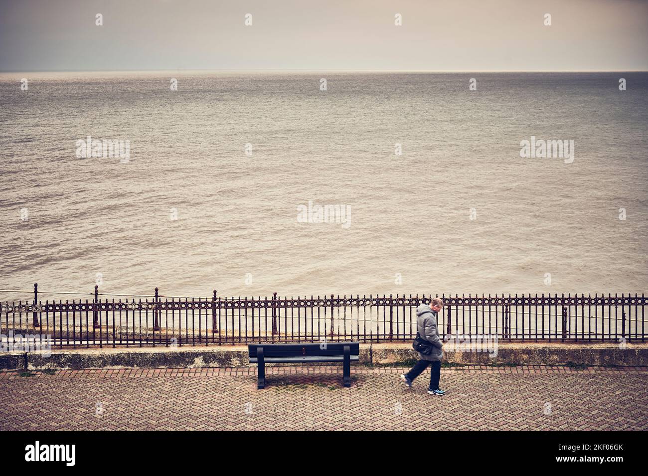 Uomo che cammina davanti a una panca vuota di fronte al mare aperto in inverno Foto Stock