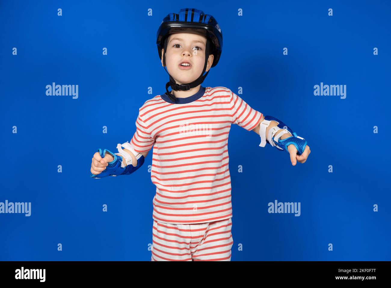 Un ragazzo felice in un casco protettivo, cuscini per gomito e guanti su sfondo blu. Protezione quando si guida una bicicletta, uno skateboard, uno skate a rotelle Foto Stock