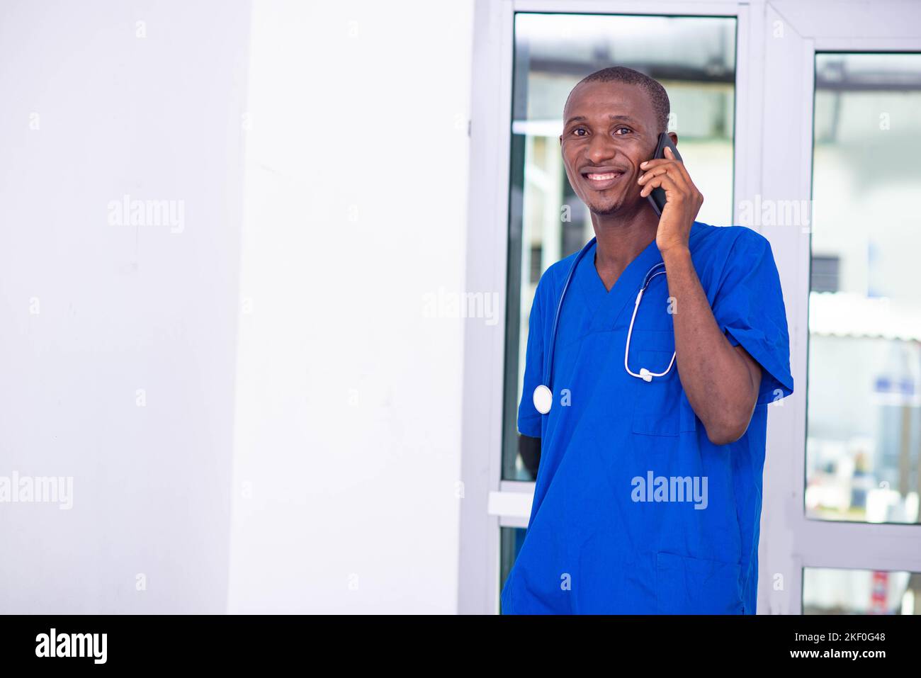 bel giovane medico maschio in uniforme blu con stetoscopio che parla sul telefono cellulare in ospedale mentre sorride. Foto Stock