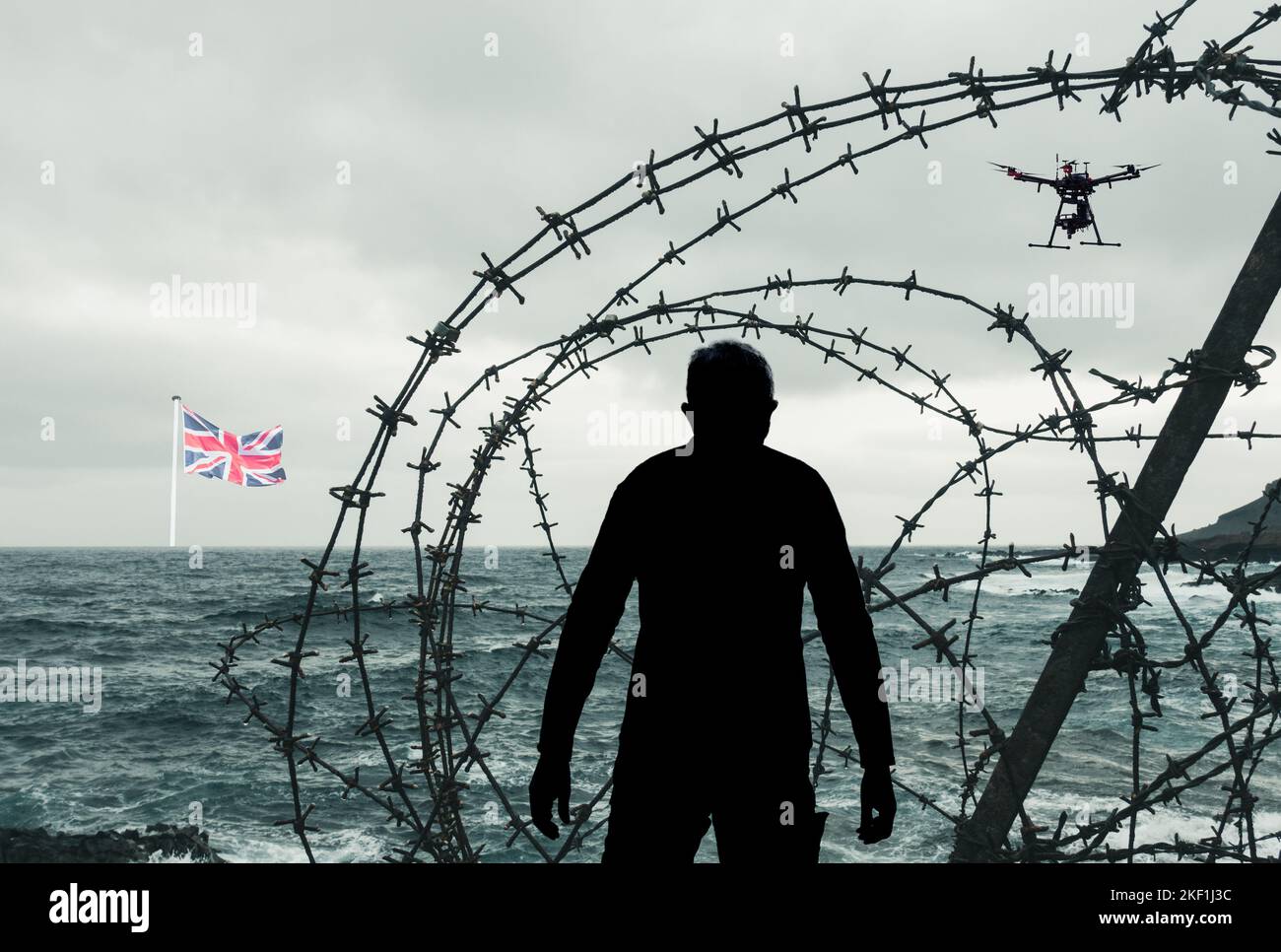Uomo che guarda attraverso recinzione spinato filo sulla spiaggia con bandiera del Regno Unito in distanza e drone overhead. Francia Regno Unito controllo di frontiera, migranti, asilo... Foto Stock