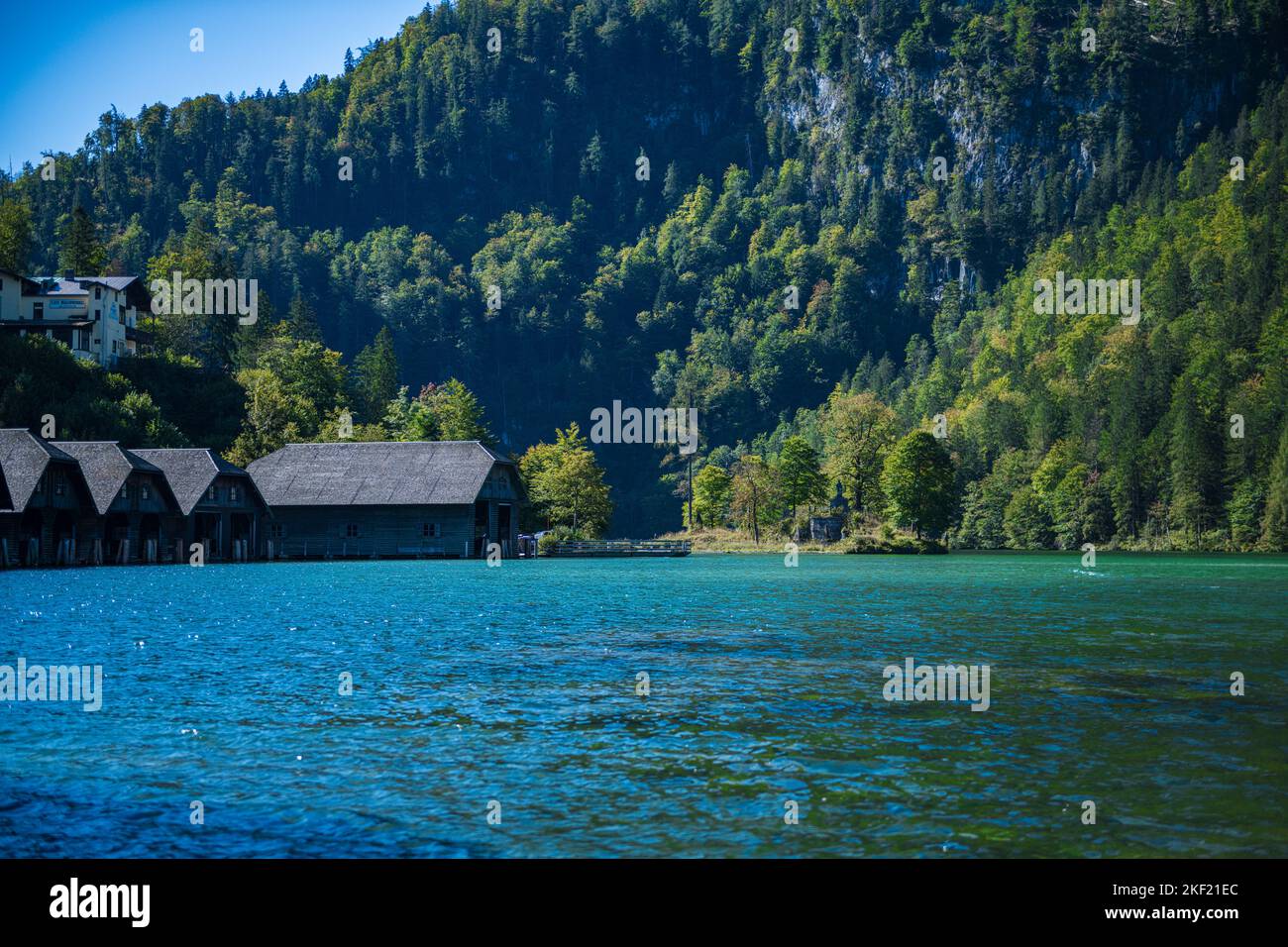 Una vista impressionante sul lago verde smeraldo Konigssee in Germania, circondato da scogliere rocciose boscose, con cottage sullo sfondo, in una giornata di sole Foto Stock