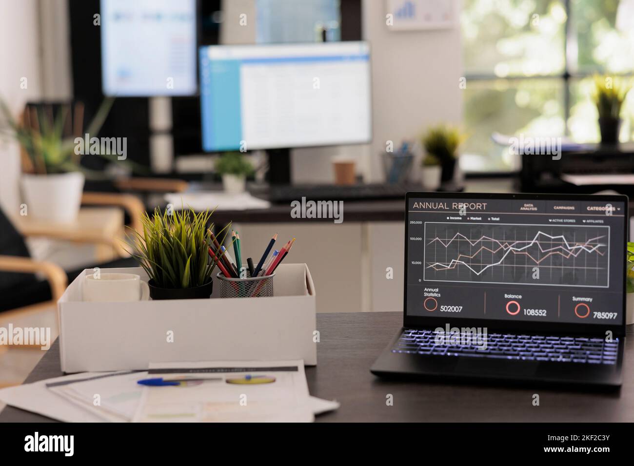 Società di analisi finanziaria con postazioni di lavoro vuote prima dell'arrivo del personale la mattina presto. Computer portatile sulla scrivania con rapporto annuale sulle vendite, schermi elettronici in background. Foto Stock