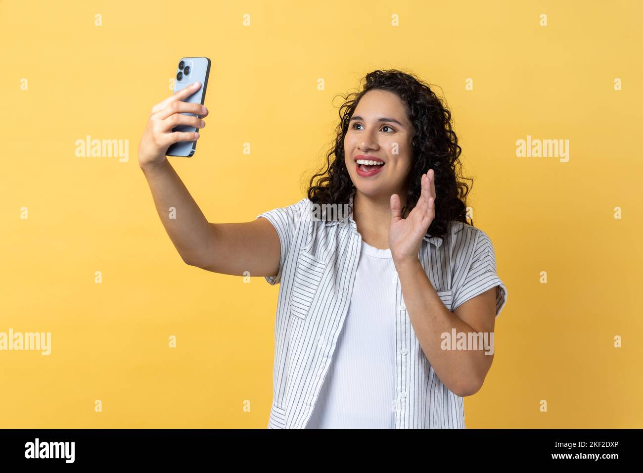 Ritratto di donna amichevole con capelli scuri ondosi tenere smartphone facendo selfie o videochiamata, ondeggiante il saluto gesto con la mano. Studio al coperto isolato su sfondo giallo. Foto Stock