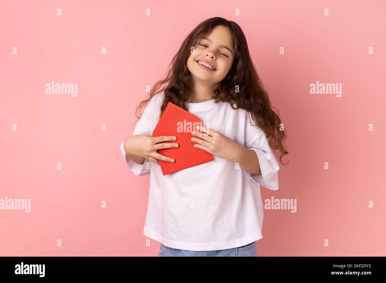 Ritratto di piccola ragazza soddisfatto che indossa T-shirt bianca in piedi aperta busta rossa con congratulazioni, lettura lettera romantica, essendo toccato. Studio in interni isolato su sfondo rosa. Foto Stock