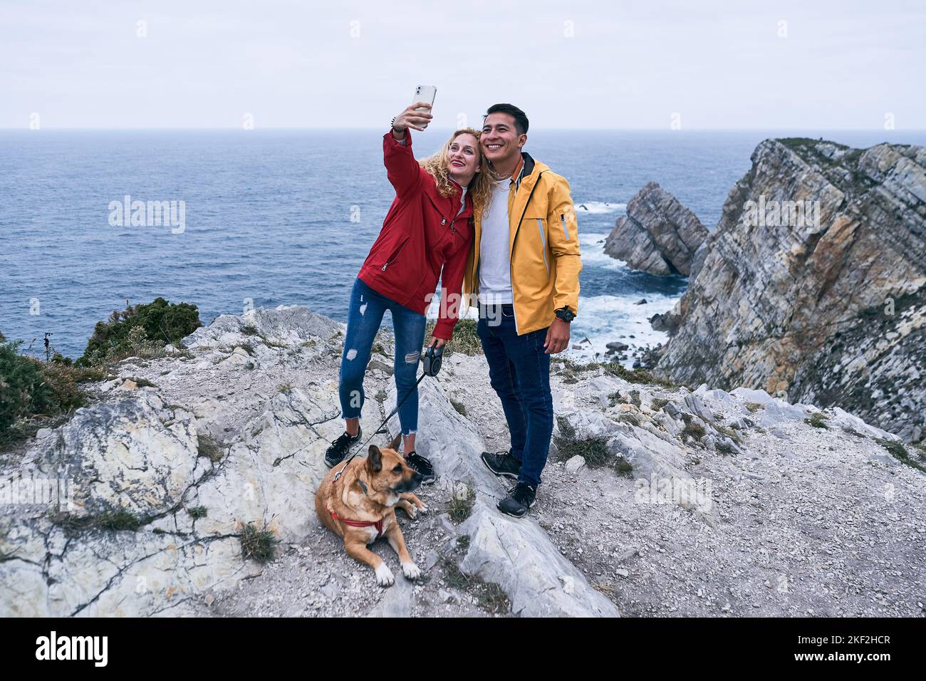 ragazza caucasica bionda del ragazzo latino e un grande cane sdraiato sorridendo mentre la ragazza prende un selfie con il suo smrtphone con la schiena al mare, cabo de Foto Stock