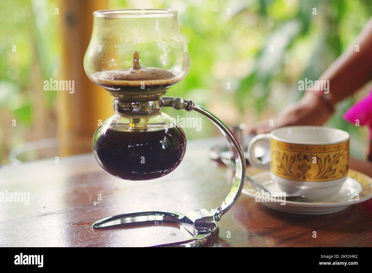 Un bollitore per caffe' a sifone di vetro; sullo sfondo, una tazza da caffe' con un tradizionale motivo batik Balinese in una piantagione di te' e caffe' a Ubud, Bali Foto Stock