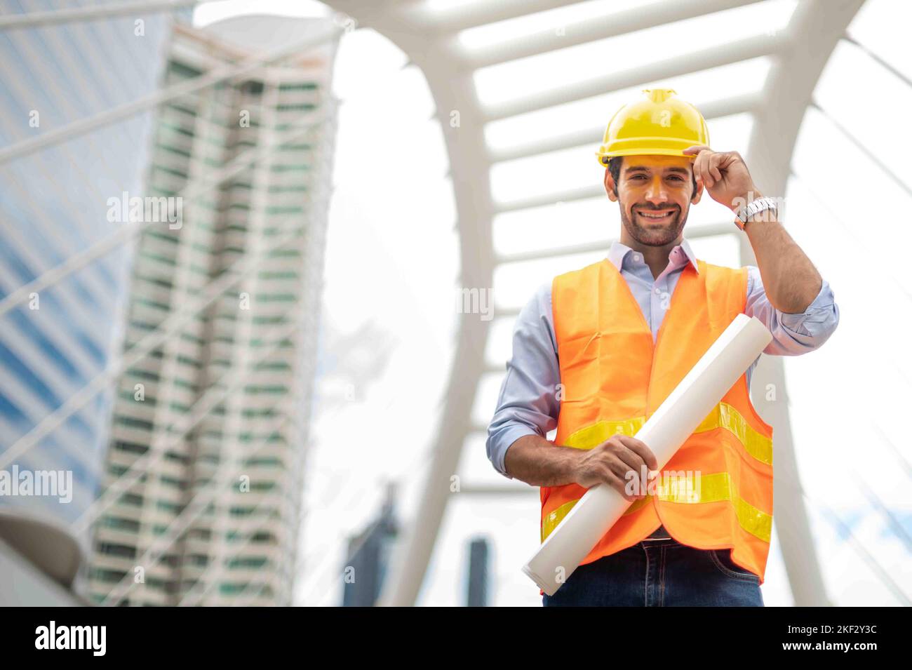 Giovane uomo caucasico con un grande foglio di carta, un ragazzo che indossa una camicia blu chiaro e jeans con un gilet arancione e casco giallo per la sicurezza in un'area di costruzione. Foto Stock