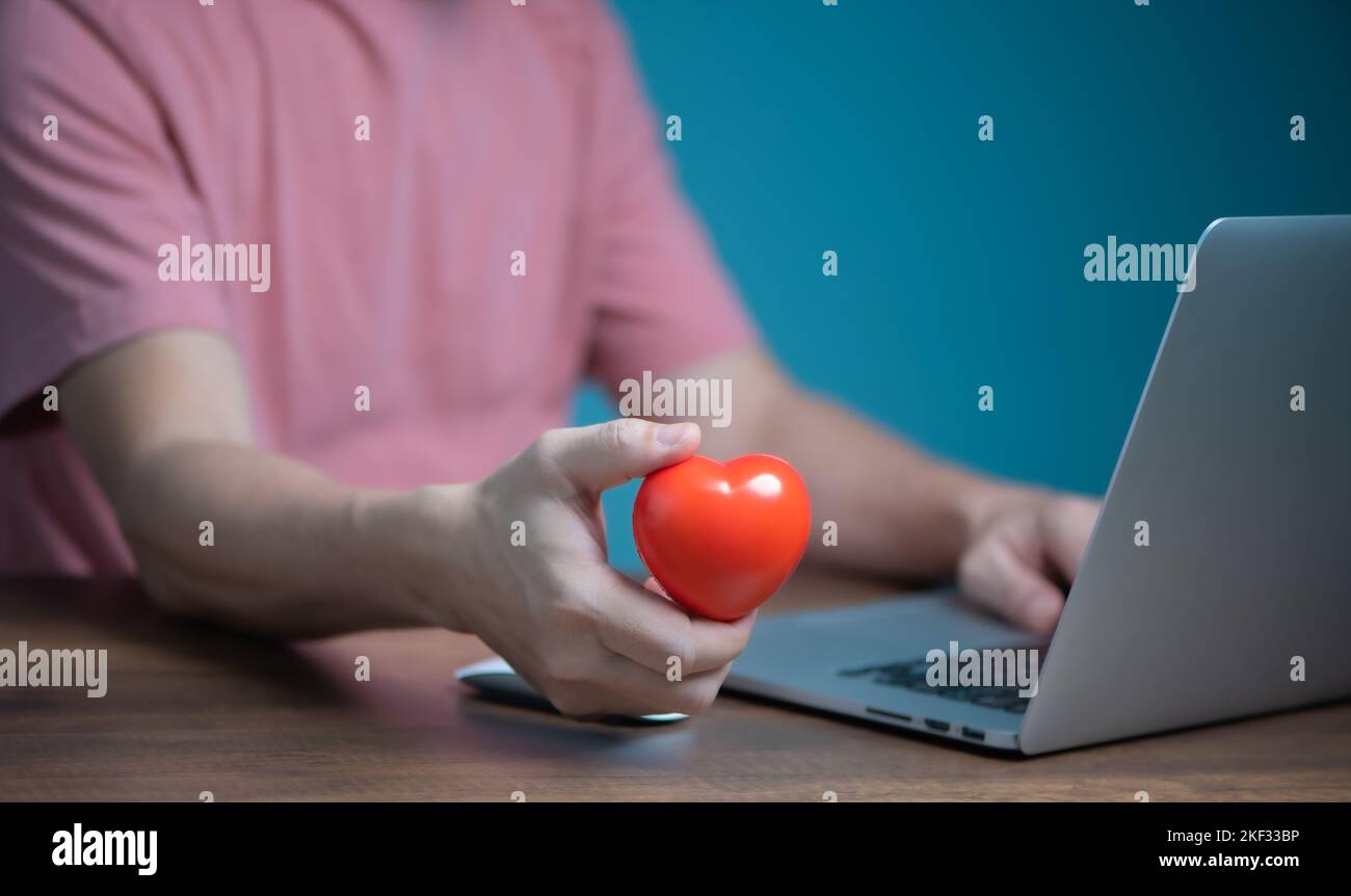 Mano dell'uomo che tiene il cuore rosso ed usando il calcolatore del laptop, dando, donazione, assicurazione, chiacchierata d'amore il concetto in linea di datazione. Foto Stock