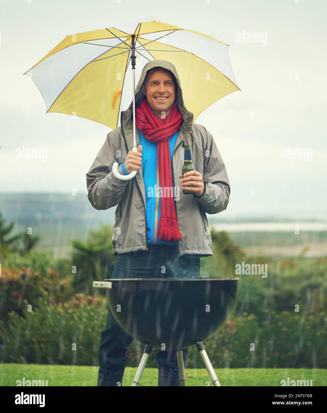 Abbiamo il coraggio, non importa il tempo: Un uomo allegro che barbecue sotto la pioggia mentre tiene un ombrello e beve una birra. Foto Stock