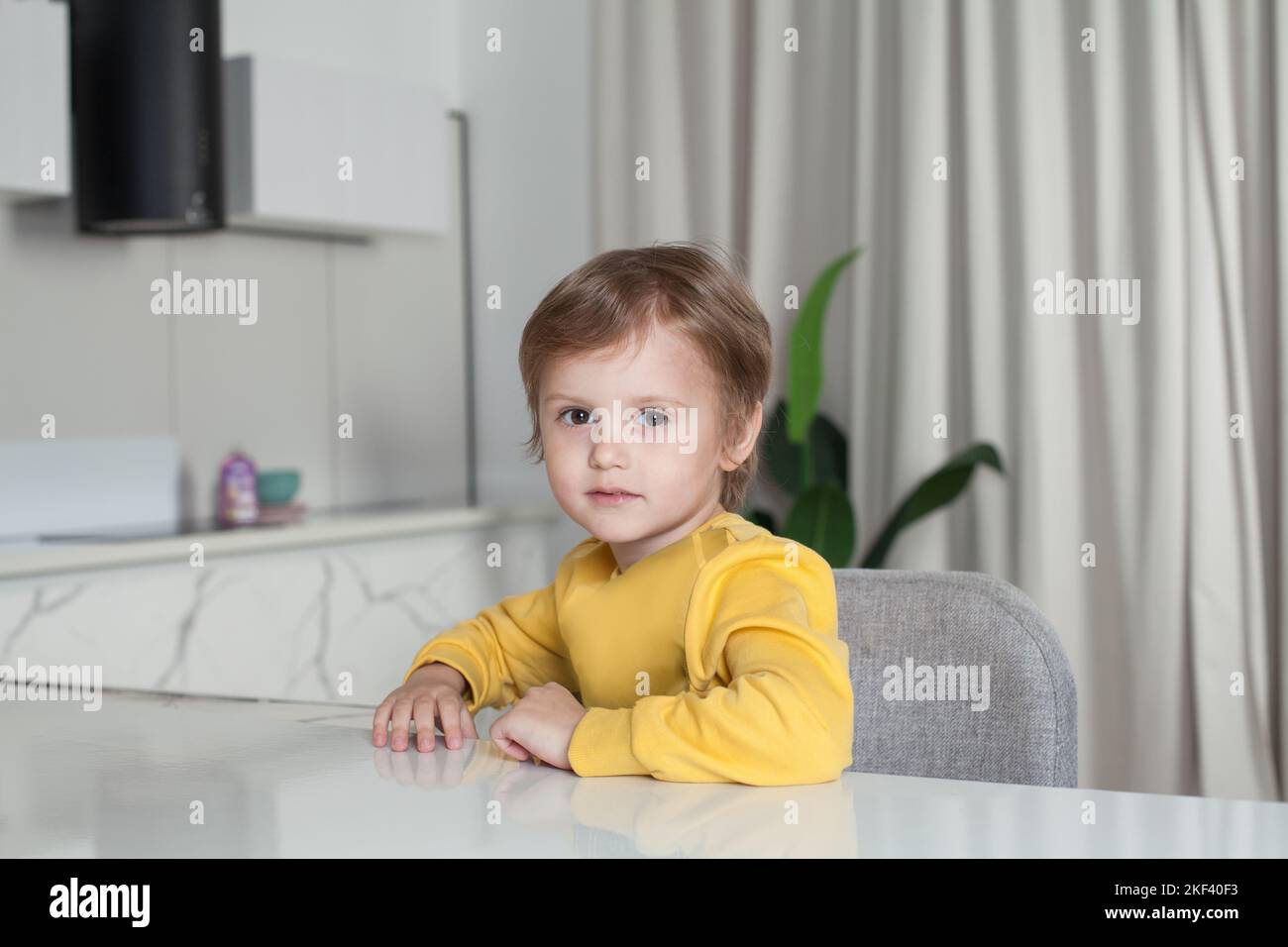 Ragazzo felice con i capelli castani in maglione giallo seduto sullo sfondo della cucina del monolocale Foto Stock